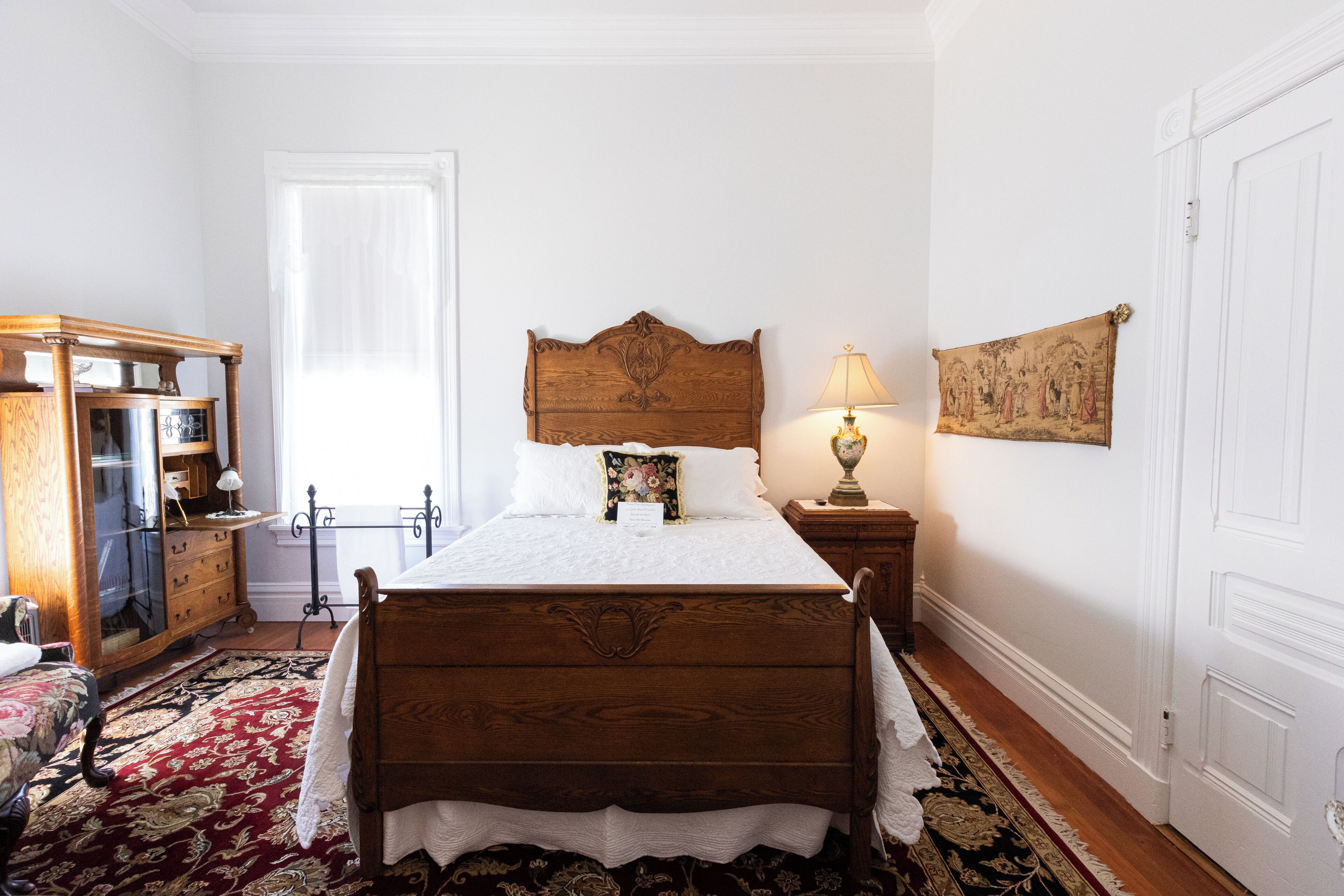 A bedroom with a wooden bed, a nightstand with a lamp, a dresser, and a large window.