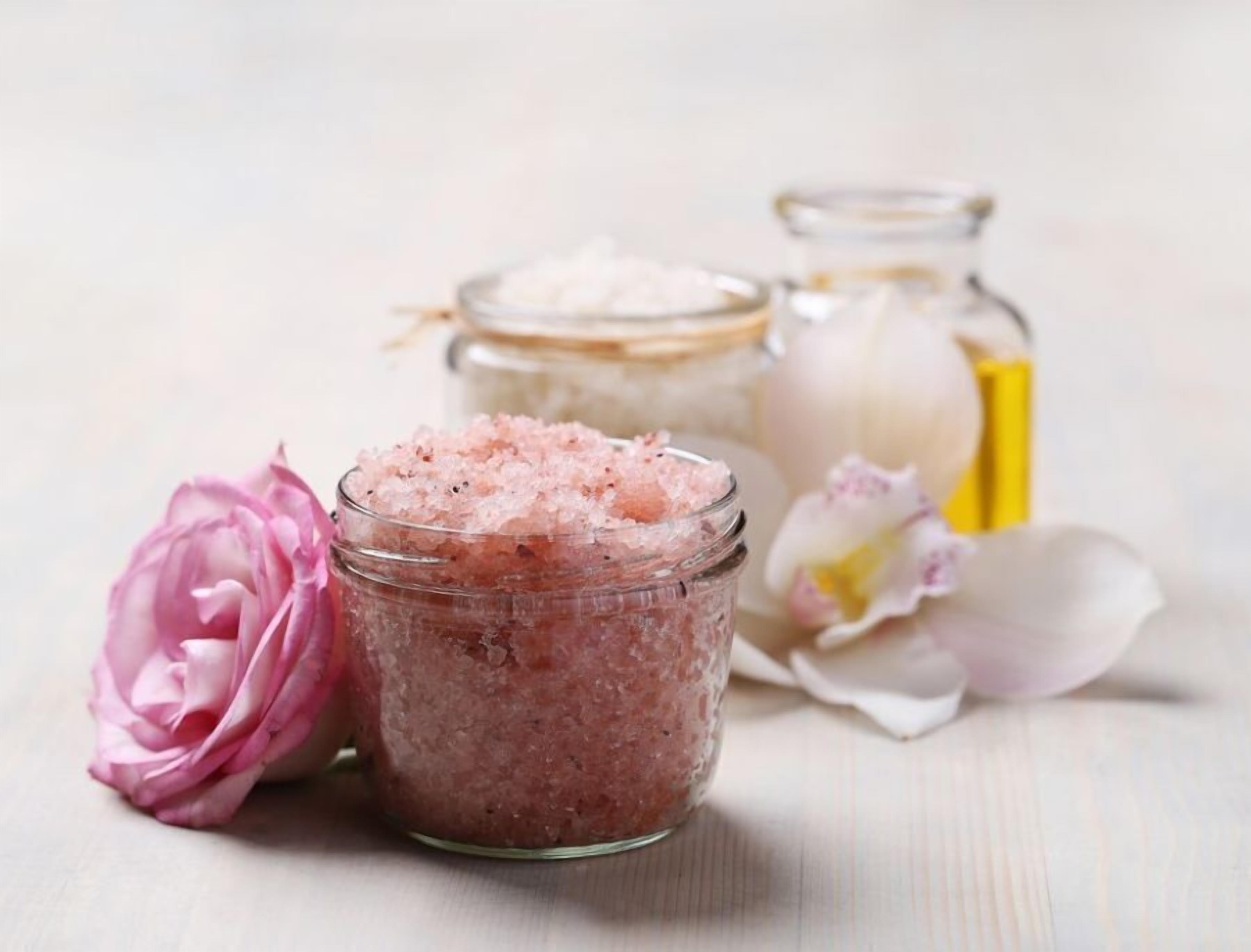 A glass jar filled with pink bath salts, a pink flower, and two small glass bottles with white and yellow contents.