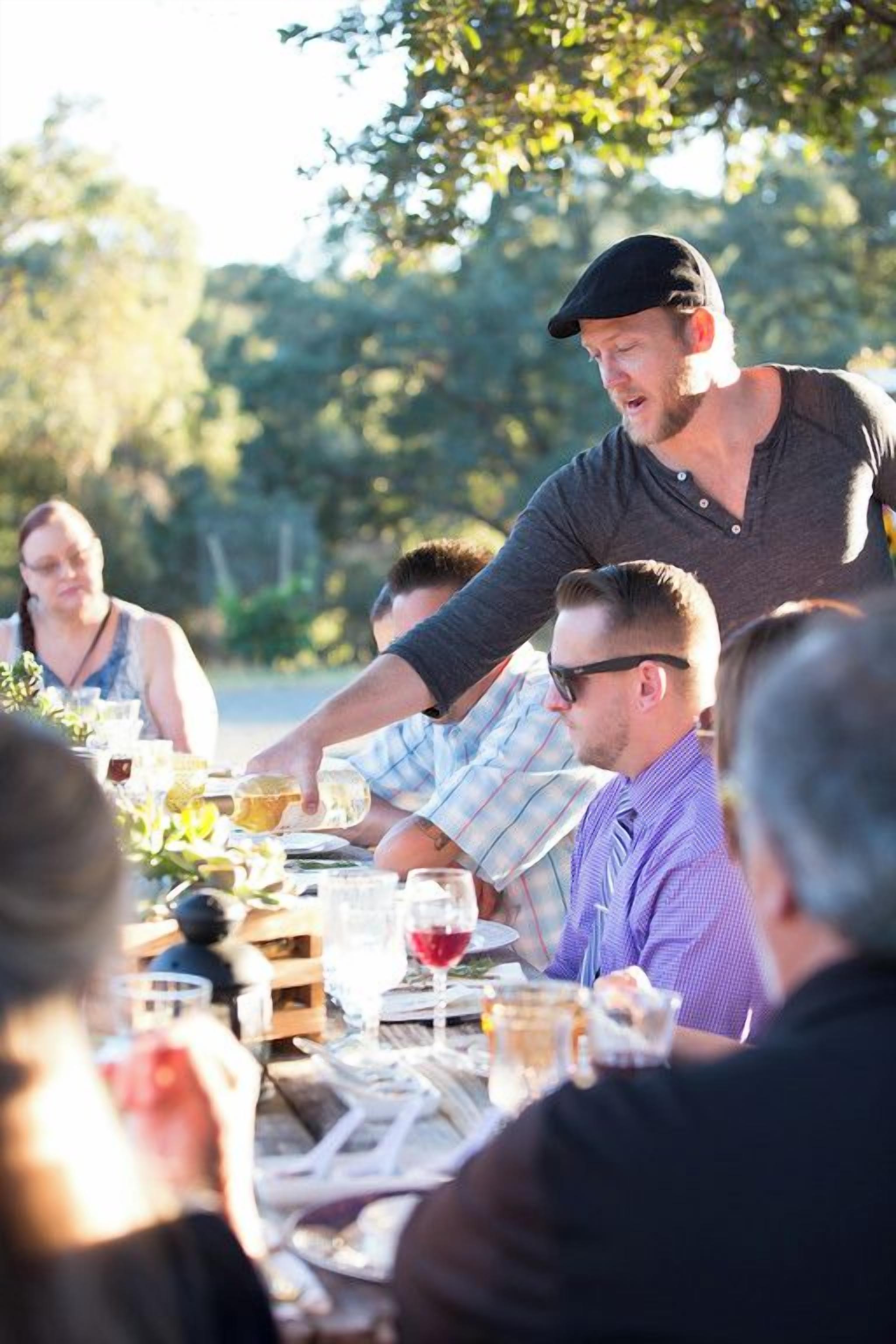 A group of people dining outdoors at a wooden table with various dishes and drinks.