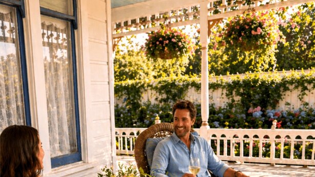 En inglasad veranda med möbler i rotting, ett bord med en vas med blommor och ett vitt trästaket.