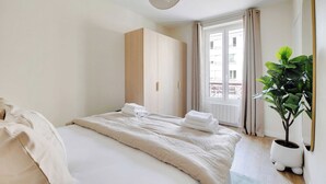 Un lit bien fait, avec des serviettes à portée de main, une grande fenêtre avec des rideaux, une plante en pot et une armoire.