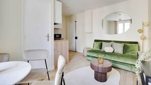 Un salon moderne avec un canapé vert, un miroir rond et une porte blanche donnant sur une autre pièce.