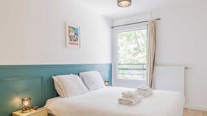 Una cama bien hecha con ropa de cama y toallas blancas, una mesita de noche con una lámpara, un cuadro en la pared y una ventana con vistas a árboles.