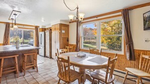 Eine Küche mit Holzschränken, einer zentralen Kochinsel mit Barhocker, einem Esstisch mit Stühlen und einem großen Fenster mit Vorhängen.