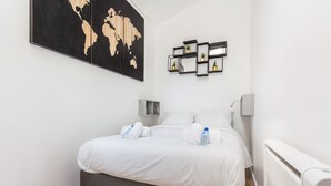 A neatly made bed with white linens and towels, a world map on the wall, and a series of black-framed shelves with potted plants.