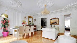 A spacious room with a wooden herringbone floor, white walls, and a large chandelier. There is a dining area with a table and chairs, and a white sofa with ottomans.