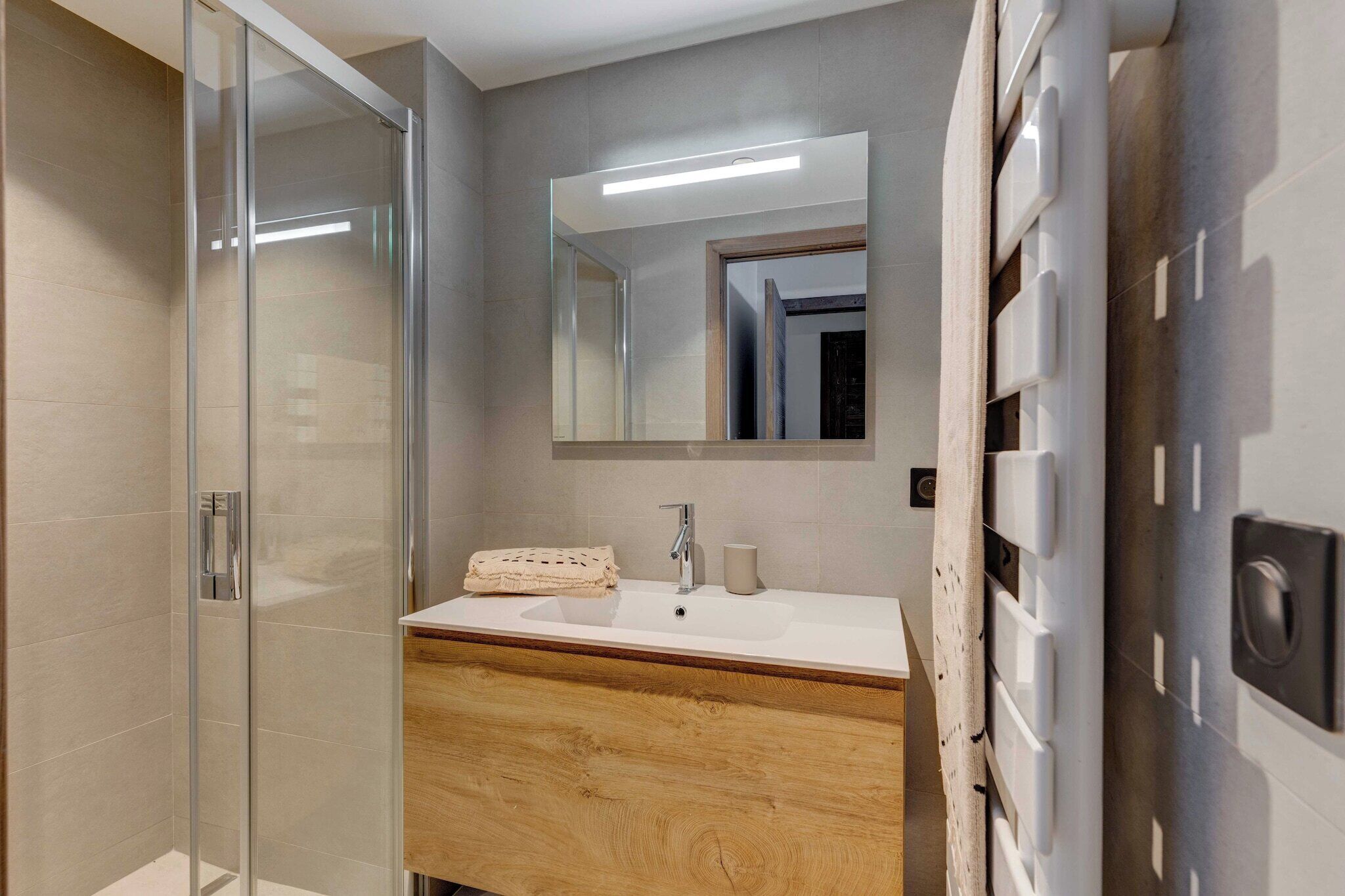 A modern bathroom with a glass shower enclosure, a wooden vanity, a large mirror, and a built-in heater.