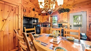 Interior de una cabaña de madera con una mesa de comedor dispuesta para cuatro personas, rodeada de sillas de madera. Un candelabro cuelga del techo y se observa una ventana con puerta de vidrio decorativa.
