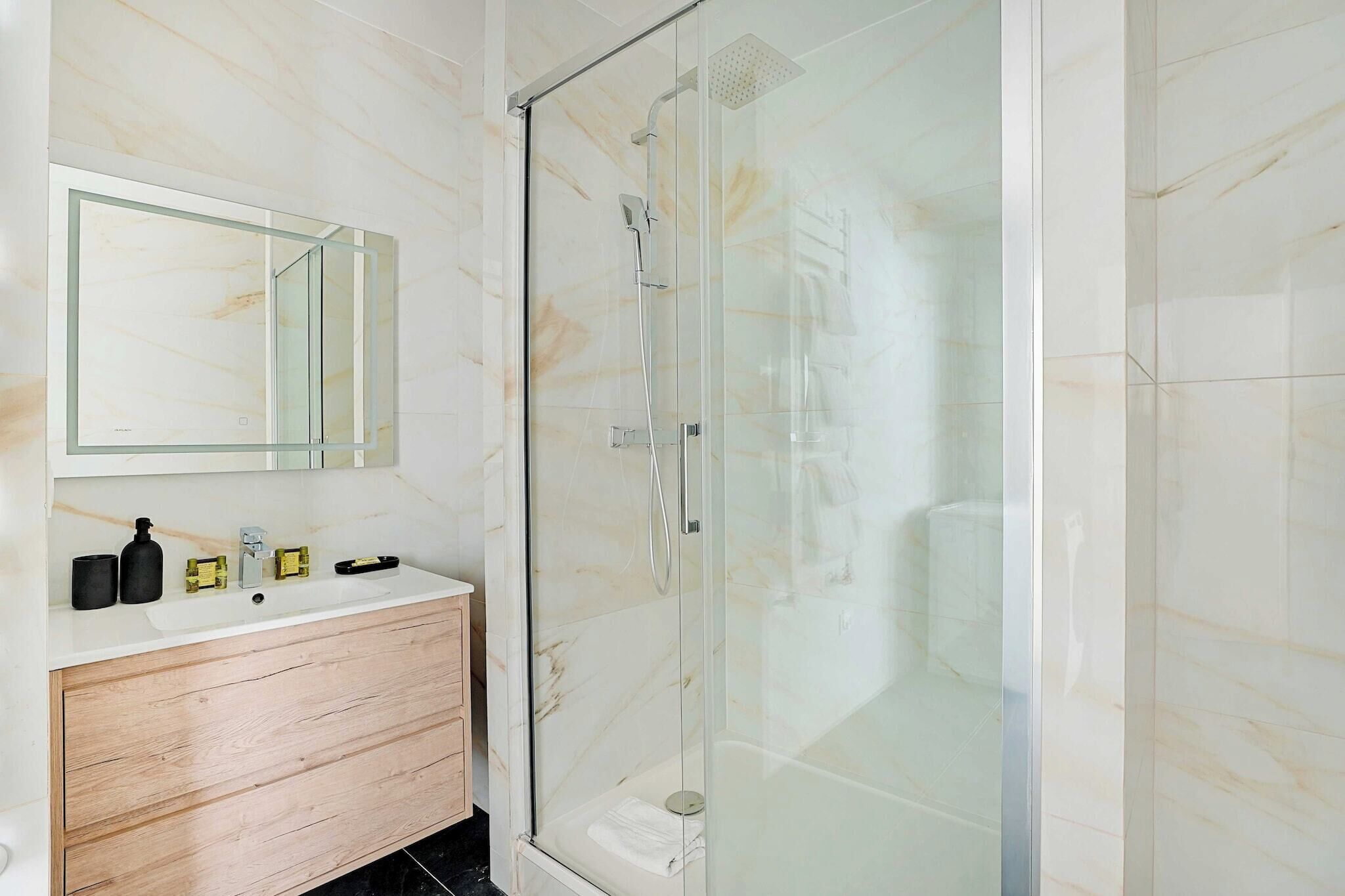 A modern bathroom with a glass-enclosed shower, a marble-tiled wall, and a wooden vanity with a mirror.