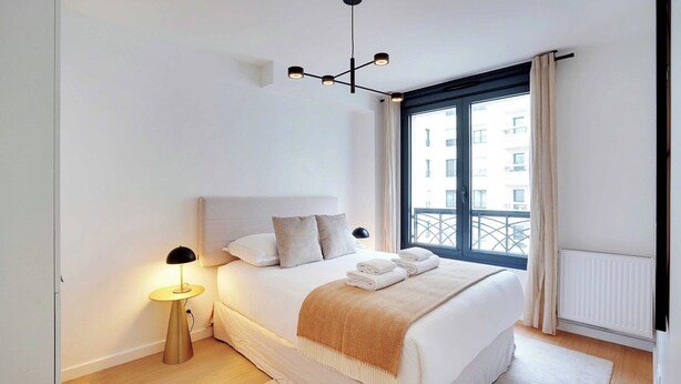 Un dormitorio moderno con una cama grande, una lámpara de noche, vista de la ciudad a través de una ventana y un radiador.