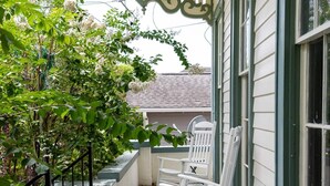 Une véranda avec une balancelle blanche et un banc, un petit arbre aux fleurs blanches et une rambarde en métal noir.