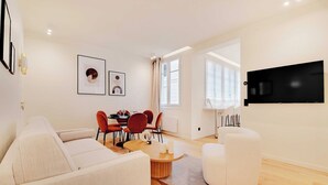 A modern living room with a sofa, a dining table with chairs, a flat-screen TV mounted on the wall, and a large window providing natural light.