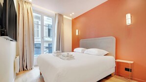 A hotel room with a large bed, a TV, a small table, and a balcony with curtains.
