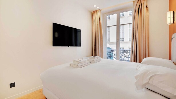 A hotel room with a bed, a flat-screen TV mounted on the wall, and a window with curtains.