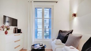 A modern hotel room with a sofa, a coffee table, a flat-screen TV, and a window with a view of buildings.