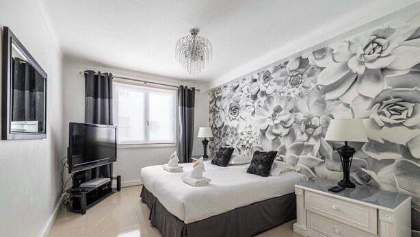 A modern bedroom with a large bed, a flat-screen TV, a chandelier, and a wall mural.