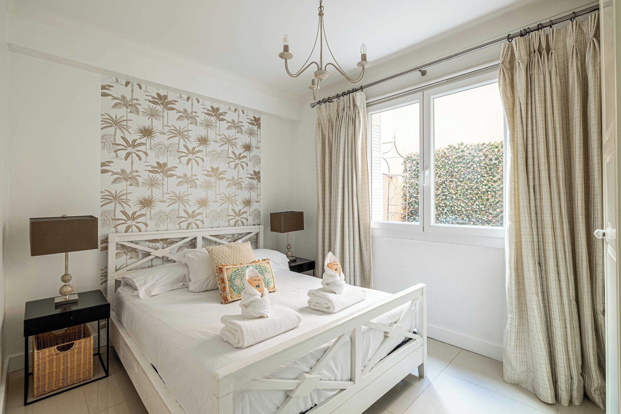 Een slaapkamer met een bed, twee nachtkastjes, een kroonluchter en een raam met gordijnen.