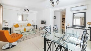 A modern living room with a glass dining table, a sofa with yellow cushions, a glass coffee table, and a chandelier.