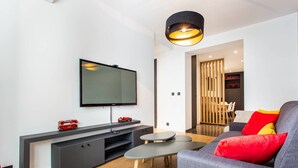 Una sala de estar moderna con un televisor de pantalla plana, un sofá gris con cojines rojos y amarillos, una mesita redonda y un aparador negro con un autobús de juguete rojo.