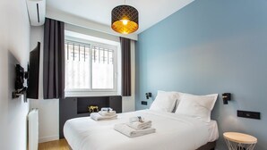 Una habitación de hotel moderna con una cama grande, una mesita de noche, una mesita auxiliar redonda, una ventana con cortinas, un televisor de pared y una lámpara colgante original.