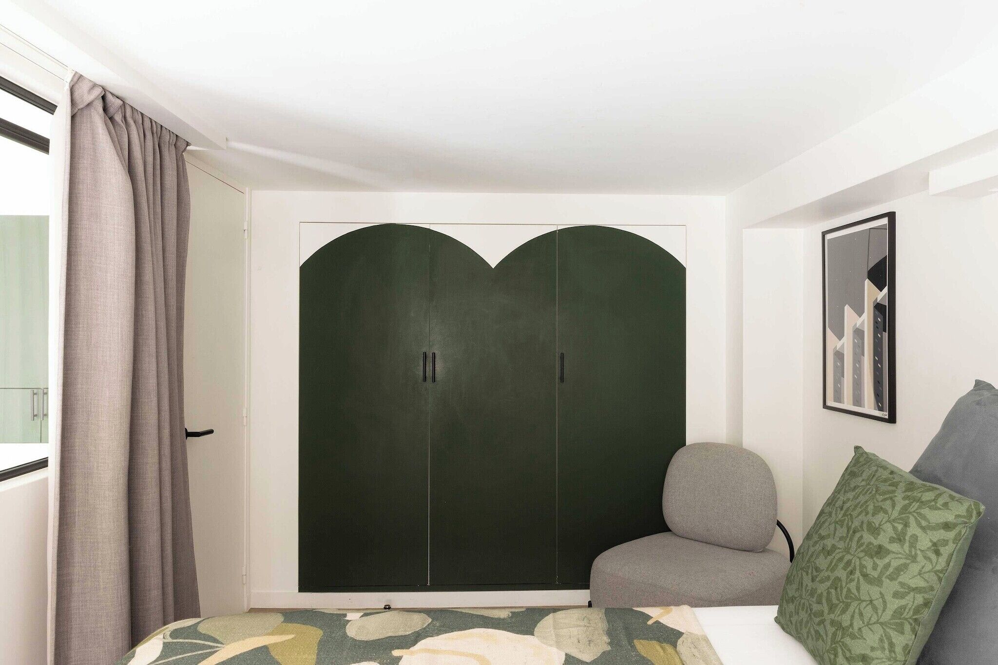 A bedroom with a large wardrobe, a grey armchair, a green pillow, and a framed picture on the wall.