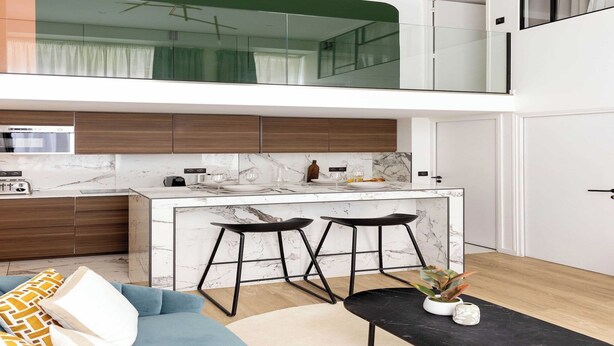 A modern kitchen with a marble countertop, two black bar stools, and a dining area with a sofa and cushions.