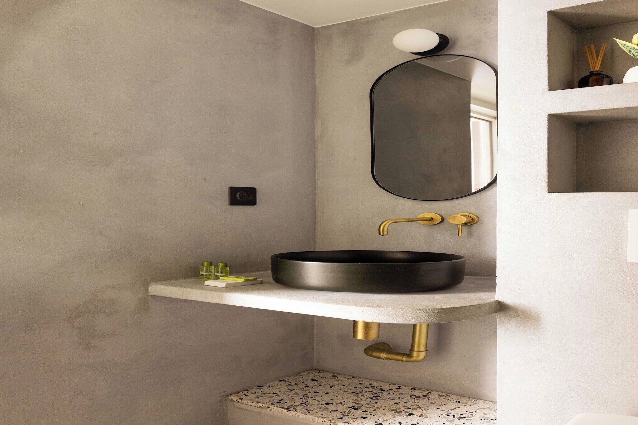 Une salle de bain moderne avec un lavabo noir rond, un comptoir blanc, un robinet doré, un miroir rond et une petite étagère contenant des articles de toilette.