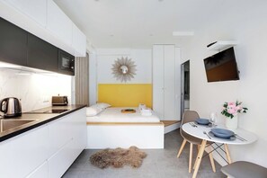 Une chambre d’hôtel compacte comprenant un coin cuisine, un lit avec une tête de lit jaune, un coin repas avec une table et des chaises, et un téléviseur fixé au mur.