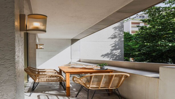 A balcony with a wooden table and wicker chairs, a potted plant, and wall-mounted lights.