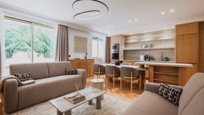 A modern living room with a sofa, coffee table, and chairs, a kitchen with wooden cabinets and appliances, and a large window with a view of trees.