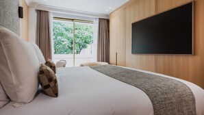 A modern hotel room with a large bed, a flat-screen TV mounted on the wall, and a view of greenery outside.