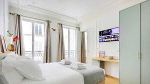 A hotel room with a bed, a TV mounted on the wall displaying a movie selection, a bookshelf with books, and a wardrobe.