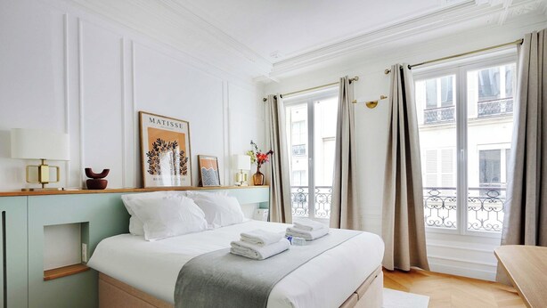 A neatly made bed with white linens and a gray throw blanket, a bedside table with a lamp, a framed artwork on the wall, and a view of the city through large windows with curtains.