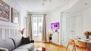 A living room with a grey sofa, wooden dining table, and chairs. There is a TV mounted on the wall, a window with curtains, and a vase with flowers on a side table.