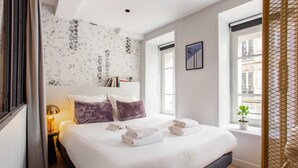 Ein ordentlich bezogenes Bett mit weißen Leinen und zwei violetten Kissen, eine Nachttischlampe und ein kleiner Tisch mit einem Buch und einer Pflanze.