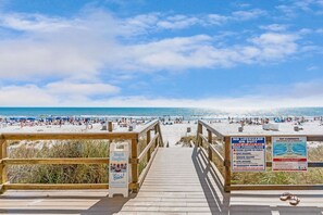 Une plage avec une promenade en bois menant au rivage, des panneaux indiquant l’absence de sauveteur et mettant en garde contre les courants de contre-rive, ainsi qu’un panneau annonçant des « feux de joie sur la plage ».