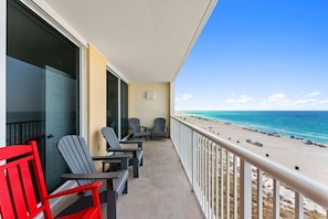 A balcony with chairs and a red chair, overlooking a beach with clear skies.