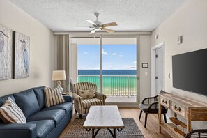 A room with a balcony, a ceiling fan, a TV, a sofa, a chair, and a table.