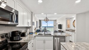 Une cuisine moderne avec des armoires blanches, des appareils en acier inoxydable, un grand îlot central et une vue sur l’extérieur.