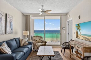 Una habitación con vista al océano, un ventilador de techo, un sofá, una silla, una mesa y un televisor.