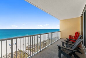 A balcony with a view of the beach and ocean, a railing, and two Adirondack chairs.
