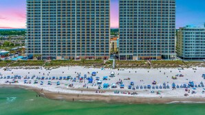 Um hotel na praia com vários edifícios altos, uma praia arenosa com diversos guarda-sóis e vista clara para o oceano.