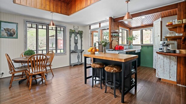 A rustic kitchen with wooden beams, a wooden dining table with chairs, and a bar area with stools. - Bellbird Rise Off-Grid Cottage (Conondale)