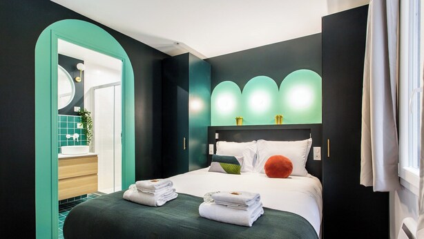 A modern hotel room with a bed, a bathroom with a mirror, and a green wall.