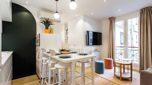 A modern kitchen with white appliances, a dining area with white chairs, a TV mounted on the wall, and a balcony with a view of the city.