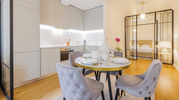 Una cucina moderna con mobili bianchi, una zona pranzo con un tavolo rotondo e sedie rivestite di grigio, e una camera da letto con un letto e un pannello di vetro.
