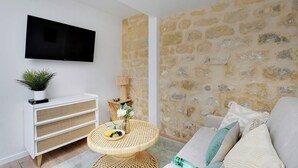 Ein modernes Wohnzimmer mit Steinwand, Flachbildfernseher, weißem Lowboard, rundem Rattantisch mit Früchten und grauem Sofa.