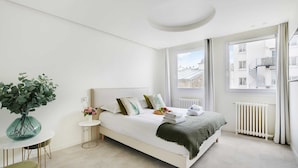 A modern bedroom with a large bed, a small round table with a vase, and a view of the city through large windows.