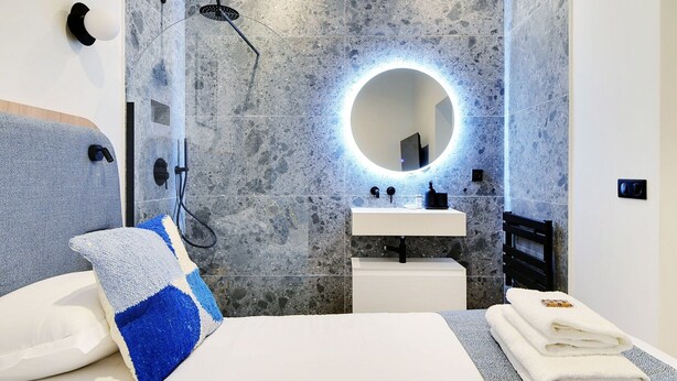 A modern bathroom with a built-in shower, a round mirror, a white sink, and a bed with towels.