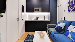 A modern kitchen with dark cabinets, a microwave, and a sink. There is a blue sofa with patterned cushions and a white vase on a coffee table.